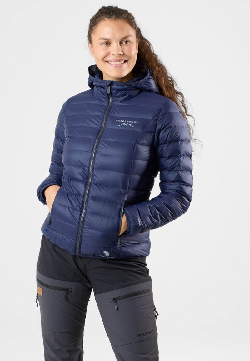 Navy insulated jacket with a zip front and hood, featuring a quilted design and logo detail, paired with dark gray pants with zippered pockets.