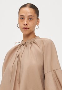 Beige blouse with a soft, lightweight fabric, featuring a round neckline, drawstring detail, and bell-shaped sleeves.