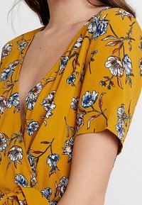 Květinové šaty s volným střihem v hořčicově žluté látce s modro-bílými květinovými vzory. Mají krátké rukávy a zavazování v pase.