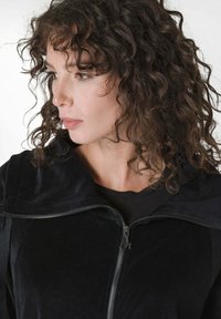 Giacca nera in velluto con zip e grande cappuccio, caratterizzata da una texture liscia e una zip argentata. Il modello mostra un profilo laterale.