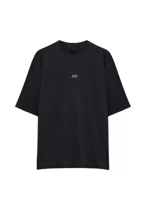 Giovane uomo con t-shirt nera Oakley e occhiali da sole futuristici grigi, che guarda oltre la spalla su sfondo bianco.