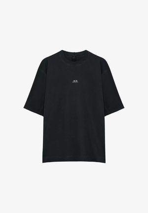 Giovane uomo con t-shirt nera Oakley e occhiali da sole futuristici grigi, che guarda oltre la spalla su sfondo bianco.