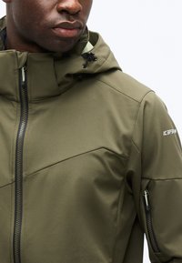 Vyras vilki alyvuogių žalios spalvos striukę su kapišonu ir juodais užtrauktukais, arti viršutinės kūno dalies ir veido dalies.