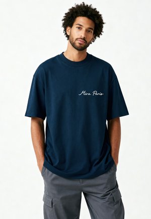 Homme aux cheveux bouclés portant un t-shirt bleu marine avec le texte « Mira Paris » et un pantalon cargo gris, debout les mains dans les poches devant un fond blanc.