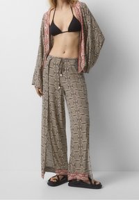 Černý bikiny top v kombinaci s širokými, vzorovanými béžovými kalhotami s červeným a bílým lemem, doplněný černými sandály. Lehké, volné provedení.