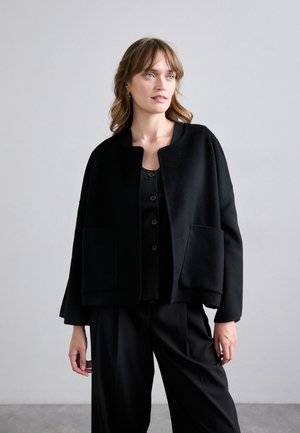 Veste en laine noire avec un col rond, deux poches avant et une coupe décontractée, portée par-dessus un haut noir à boutons et un pantalon noir.