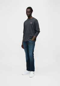 Μακρυμάνικο σκούρο γκρι πουκάμισο με ένα μικρό κόκκινο και λευκό λογότυπο, συνοδευόμενο από μπλε τζιν και λευκά αθλητικά παπούτσια. Απλός, καθημερινός σχεδιασμός.