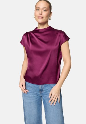 Femme aux cheveux blonds coiffés en chignon bas, portant des boucles d'oreilles créoles en or, un chemisier en satin violet à manches courtes et un jean bleu clair, debout devant un fond blanc.