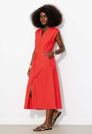 Femme avec une grande coiffure afro portant une robe midi rouge sans manches et des sandales à imprimé léopard, debout contre un mur blanc.