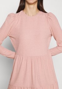 Abito a maniche lunghe di un rosa pallido realizzato in tessuto a coste. Presenta un collo rotondo, spalle arricciate e un design della gonna a strati.