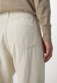 Nahaufnahme einer Person, die beige Cordhosen mit Gesäßtasche und ein hellbraunes Langarmhemd trägt, Hand ruhend an der Seite.