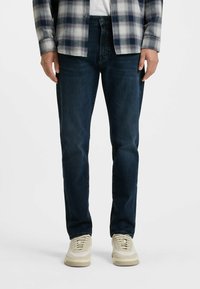 Vaqueros de mezclilla azul oscuro con un corte recto, que presentan una textura sutil. Combinados con zapatillas de color claro y una camisa a cuadros gris.