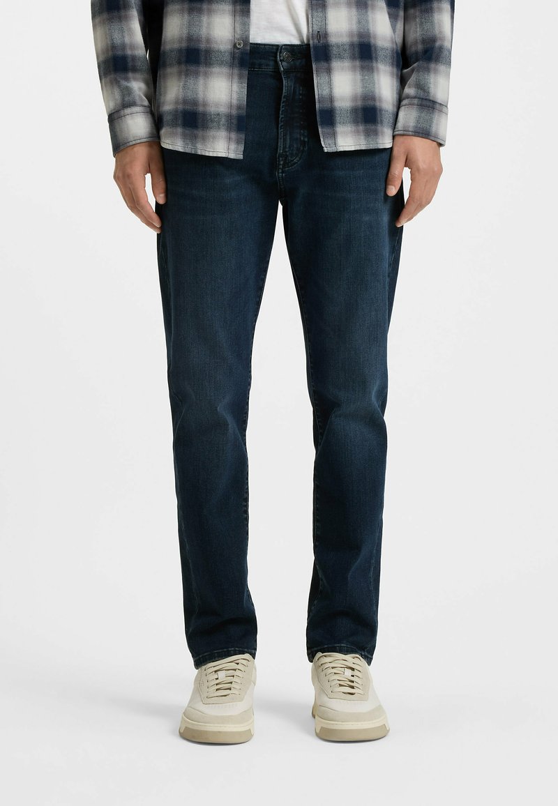 Vaqueros de mezclilla azul oscuro con un corte recto, que presentan una textura sutil. Combinados con zapatillas de color claro y una camisa a cuadros gris.