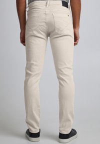 Jeans bej deschis cu croială slim, cu buzunare în spate și o plăcuță de piele neagră pe talie, asortaţi cu încălţăminte închisă la culoare.