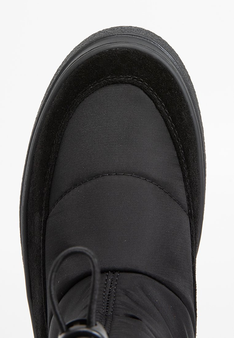 Bota de invierno negra con parte superior de nylon acolchado, acentos de gamuza, puntera redondeada y suela de goma texturada. Detalle de cordones en la parte delantera.