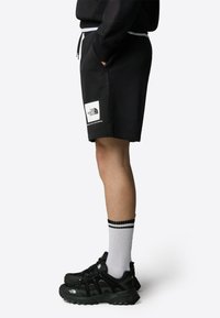 Pantalones cortos de algodón negros con una cinturilla elástica, bolsillos laterales y un parche de logo blanco. Combinados con zapatos negros y calcetines blancos con rayas negras.