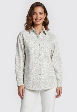 Vrouw glimlacht, draagt een lichtgrijs overhemd met dierenprint en borstzak, en een witte broek, staand tegen een effen achtergrond.