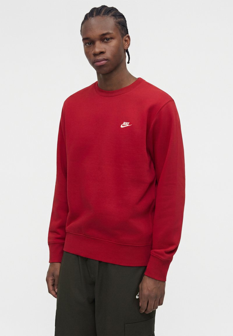 Joven con el cabello trenzado con sudadera roja de Nike y pantalones oscuros, de pie frente a un fondo claro y liso.