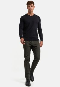 Suéter negro de cuello en V, diseño ajustado, puños acanalados, acompañado de pantalones verde oliva y zapatos de cuero negro, de pie contra un fondo blanco.