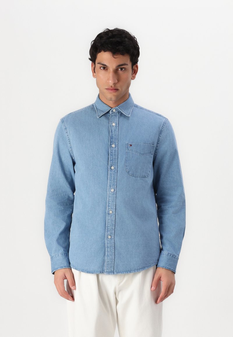 Lyseblå denimskjorte med knappeoppsett, spiss krage, en brystlomme og lett tekstur. Har en merkevarelogo på lommen.