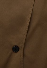 Gros plan sur un tissu brun avec un bouton noir à quatre trous cousu près du centre et une couture visible le long du bord.