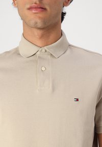Beige polo-shirt med struktureret stof, to-knaps placket og et lille broderet logo med røde, hvide og blå striber.