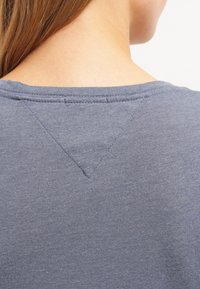Camiseta de algodón gris con cuello redondo, mangas cortas y un detalle de costura en forma de V estrecho en la parte posterior. Textura suave.