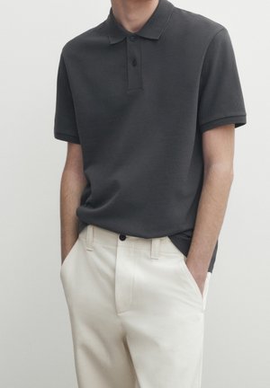 Man wearing a dark grey short-sleeve polo shirt tucked into off-white pants with hands in pockets against a plain background.