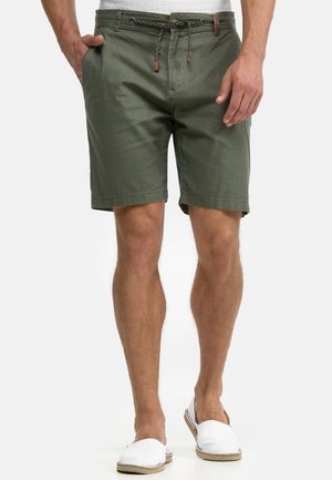 Man wearing olive green drawstring shorts and white slip-on shoes, standing with one hand in pocket against plain background.