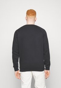 Person med kort, lockigt röd hår iklädd en svart långärmad tröja och vita byxor, stående vänd bort från kameran mot en enkel vit bakgrund.