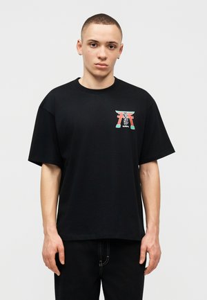 Junger Mann mit kurzen Haaren, der ein schwarzes T-Shirt trägt, auf der linken Brust mit einem kleinen, bunten Torii-Tor-Design.