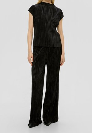 Frau trägt schwarze, plissierte Kurzarmbluse und passende plissierte, weit geschnittene Hose, steht vor einem schlichten grauen Hintergrund.