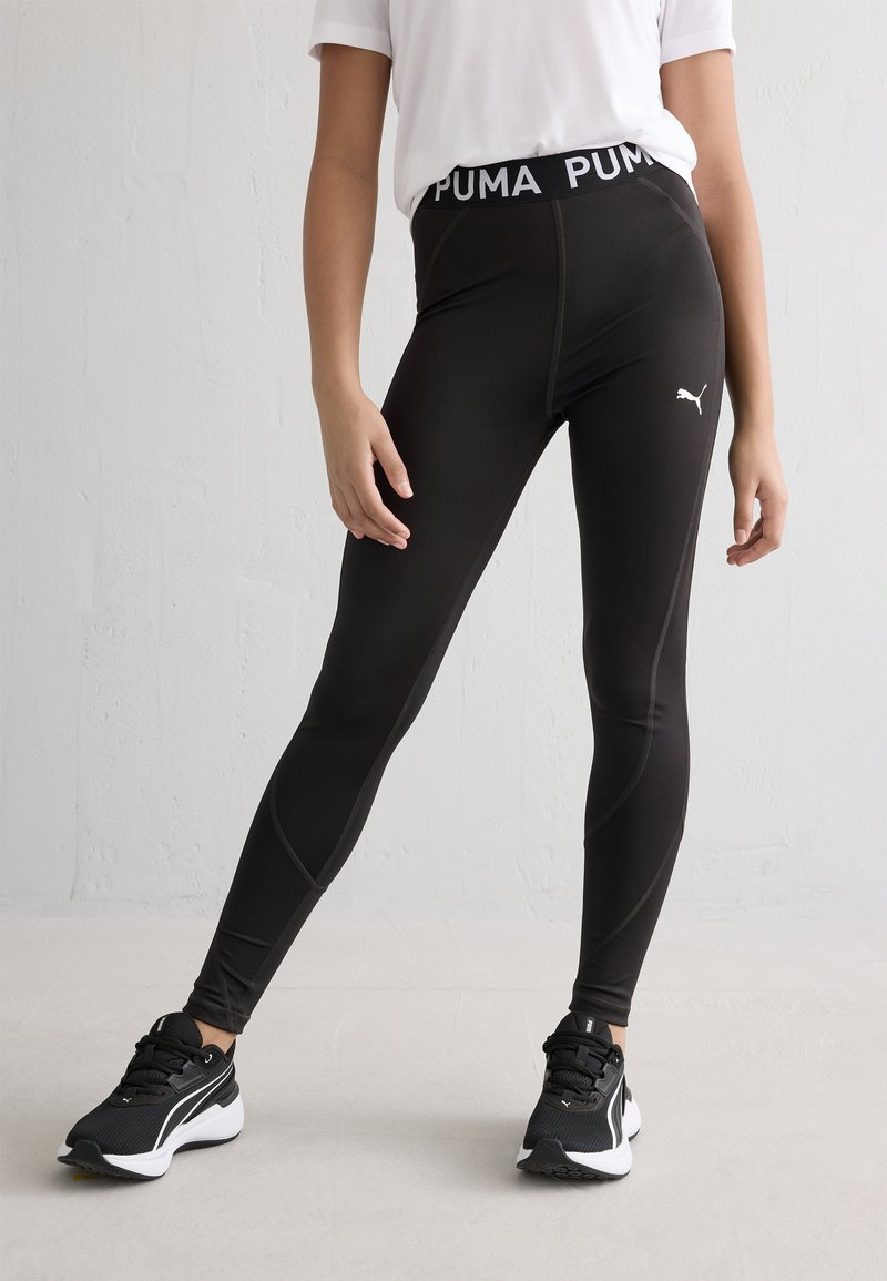 Čierne športové legíny so širokým pásom so značkou, hladká textúra, kontúrové švy, v kombinácii s čiernymi športovými topánkami s bielymi akcentmi.