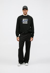 Sudadera negra con estampado gráfico, combinada con pantalones negros holgados y zapatillas negras. Los accesorios incluyen una gorra negra. Diseño sencillo.