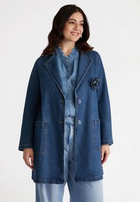 Jachetă de blugi cu o cămașă bleu deschis dedesubt. Prezintă un accent floral, buzunare mari, nasturi și o croială lejeră cu guler.