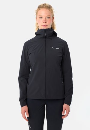 Frau mit hellen lockigen Haaren, die eine schwarze Vaude Zip-up-Kapuzenjacke und schwarze Hosen trägt und vor einem weißen Hintergrund steht.