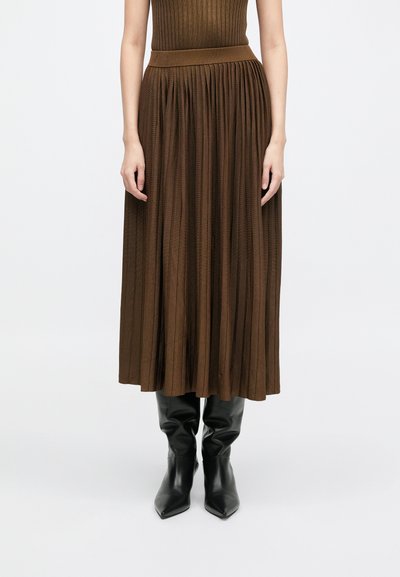Καφέ πλισέ μάξι φούστα από ριπ ύφασμα με εφαρμοστό μέσο, συνδυασμένη με μαύρες μποτίνες ψηλές μέχρι το γόνατο με μυτερή μύτη.