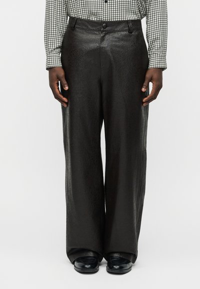 Homem vestindo calças largas de couro pretas, sapatos pretos e uma camisa branca com padrão de grade preta.