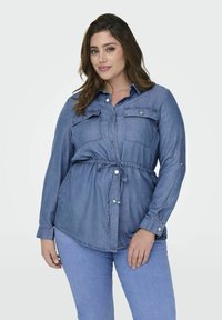 Svetlo modra denim srajca z dolgimi rokavi, gumbom spredaj, dvema žepoma na prsih in pasom z vrvico. Gladka tekstura in sproščen kroj.