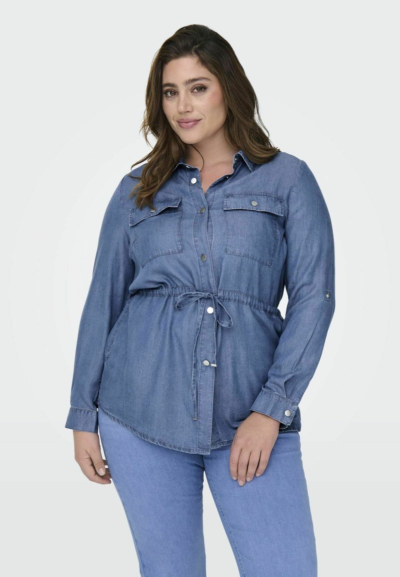 Svetlo modra denim srajca z dolgimi rokavi, gumbom spredaj, dvema žepoma na prsih in pasom z vrvico. Gladka tekstura in sproščen kroj.
