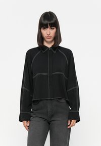 Black long-sleeve button-up shirt with contrasting white stitching, a pointed collar, and a cropped design. Paired with dark jeans.