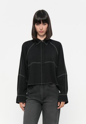 Black long-sleeve button-up shirt with contrasting white stitching, a pointed collar, and a cropped design. Paired with dark jeans.