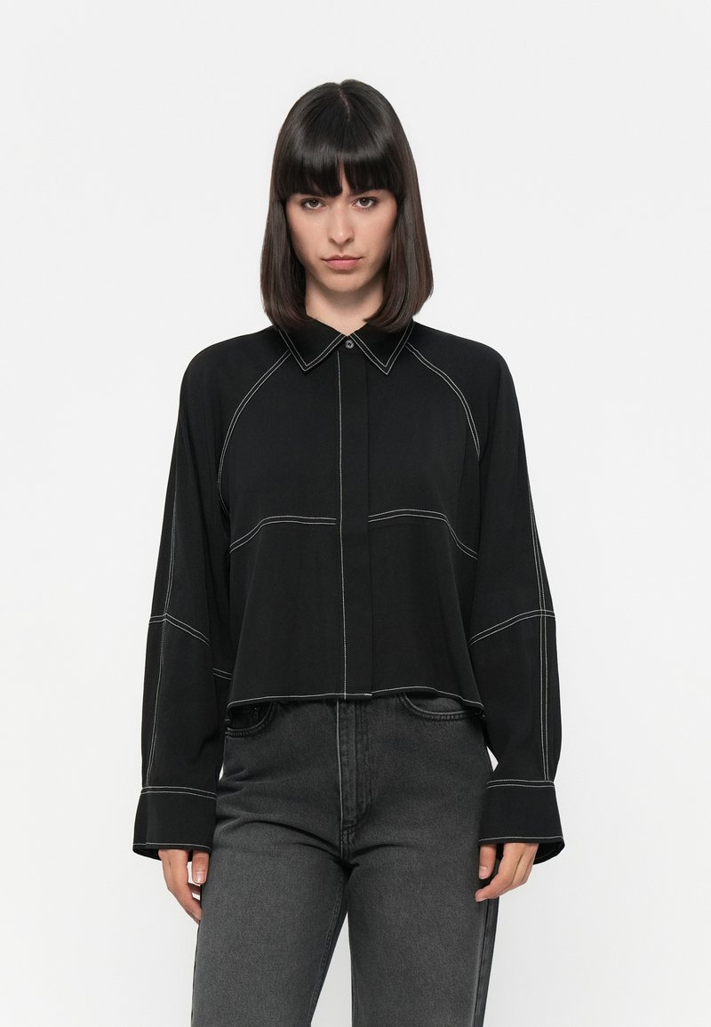 Sort langærmet skjorte med knapper, kontrasterende hvid syning, spidse krave og cropped design. Matchet med mørke jeans.