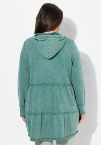 Grünes Kapuzenkleid mit langen Ärmeln aus strukturiertem Stoff, verfügt über ein mehrlagiges Design und eine Kapuze. Enthält Rüschendetails an der Taille.