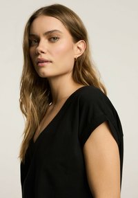 Czarny top z dekoltem w serek, krótkie rękawy, gładka faktura i luźny krój. Modelka pozuje w profilu bocznym; miękkie fale we włosach. Minimalistyczny design.