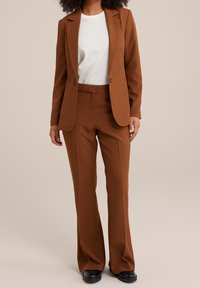 Brown tailored suit with a single-button blazer, matching pants, and a white crew-neck t-shirt. Smooth fabric and a fitted design. Black shoes.