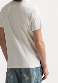 Polo shirt beige a maniche corte con una finitura liscia e colletto classico, abbinato a jeans in denim leggero e strappato di colore blu.