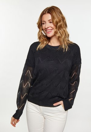 Pulover negru tricotat cu model zigzag și deschideri pe mâneci. Manșete și tiv ribbat. Purtați peste pantaloni albi.