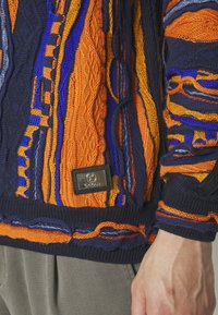 Strickpullover mit auffälligen Mustern in Orange, Blau und Marineblau. Strukturierte Zopfmuster und glatte Garnoberfläche, mit einem Metall-Label am Saum.