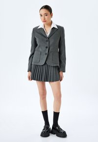 Grijze wollen blazer met een geknipt kraag en voorkeurszakken, gecombineerd met een grijze geplooide rok. Het model draagt zwarte knielange sokken en stevige schoenen.